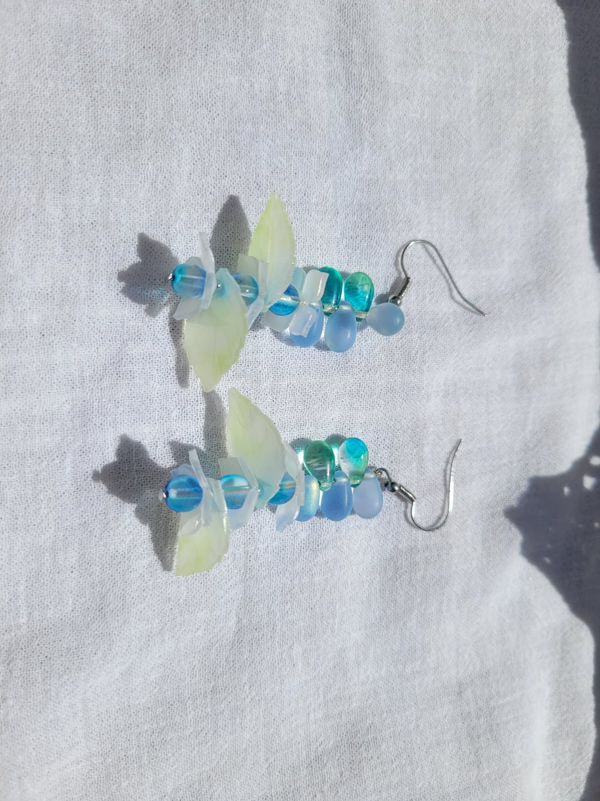 Pair of earrings with blue and green glass beads, acrylic leaves and small flowers on a white fabric background