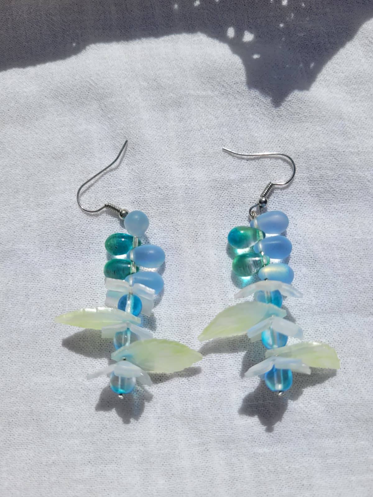 Pair of earrings with blue and green glass beads, acrylic leaves and small flowers on a white fabric background