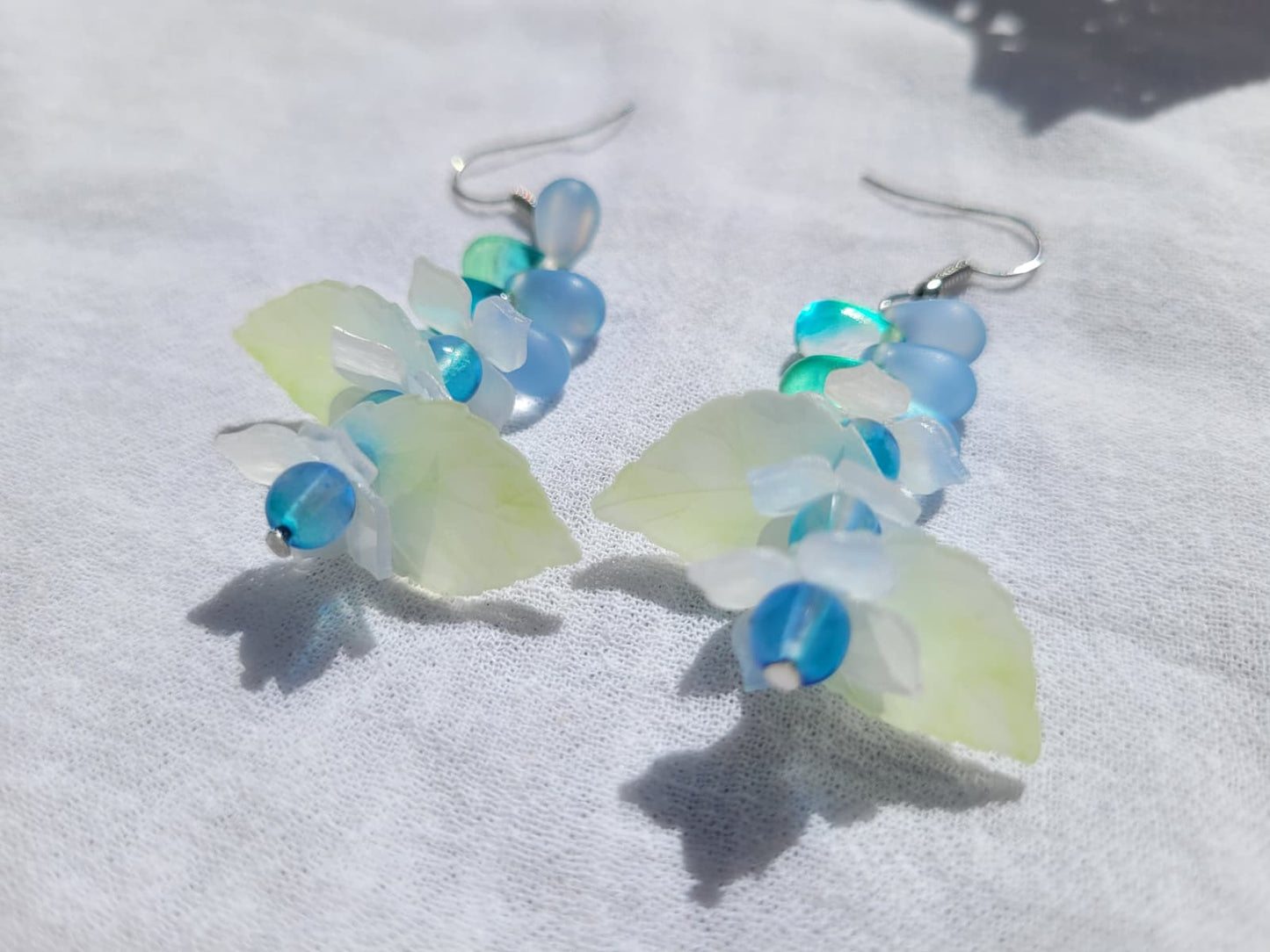 Pair of earrings with blue and green glass beads, acrylic leaves and small flowers on a white fabric background