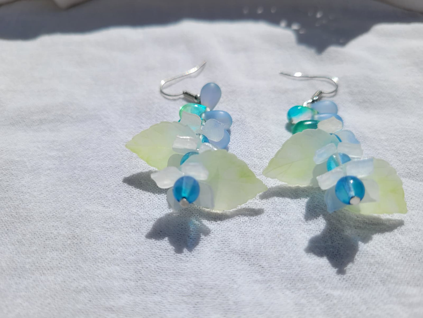 Pair of earrings with blue and green glass beads, acrylic leaves and small flowers on a white fabric background