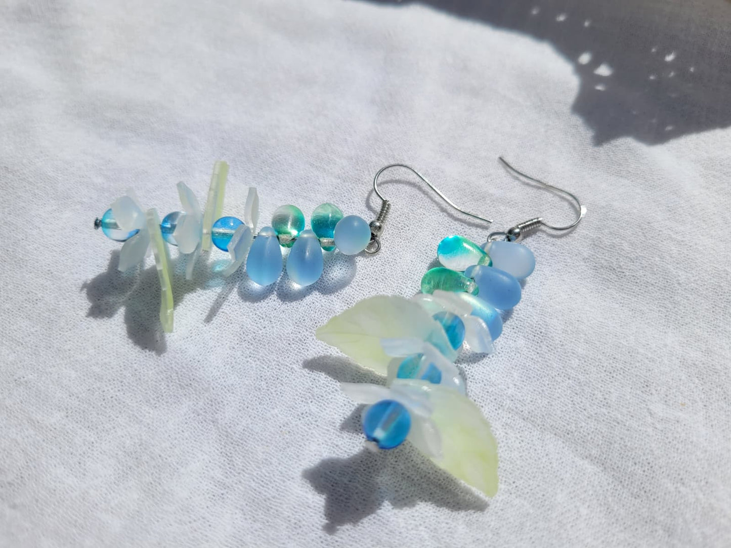Pair of earrings with blue and green glass beads, acrylic leaves and small flowers on a white fabric background