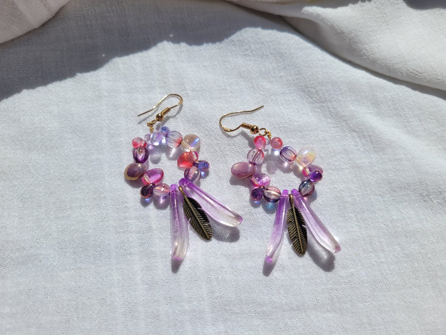 Pair of round dangle beaded earrings with pink and purple beads and bronze feather charms on a light gray background; gold-plated hooks