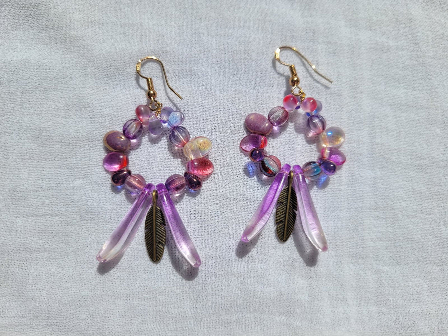 Pair of round dangle beaded earrings with pink and purple beads and bronze feather accent on a light gray background