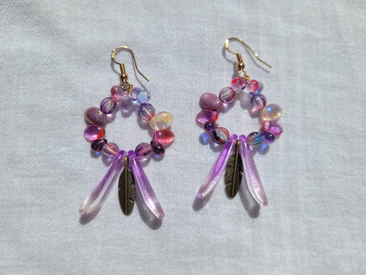Pair of round dangle beaded earrings with pink and purple beads and bronze feather accent on a light gray background
