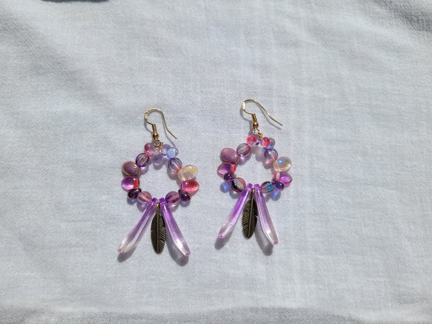 Pair of round dangle beaded earrings with pink and purple beads and bronze feather charms on a light gray background; gold-plated hooks