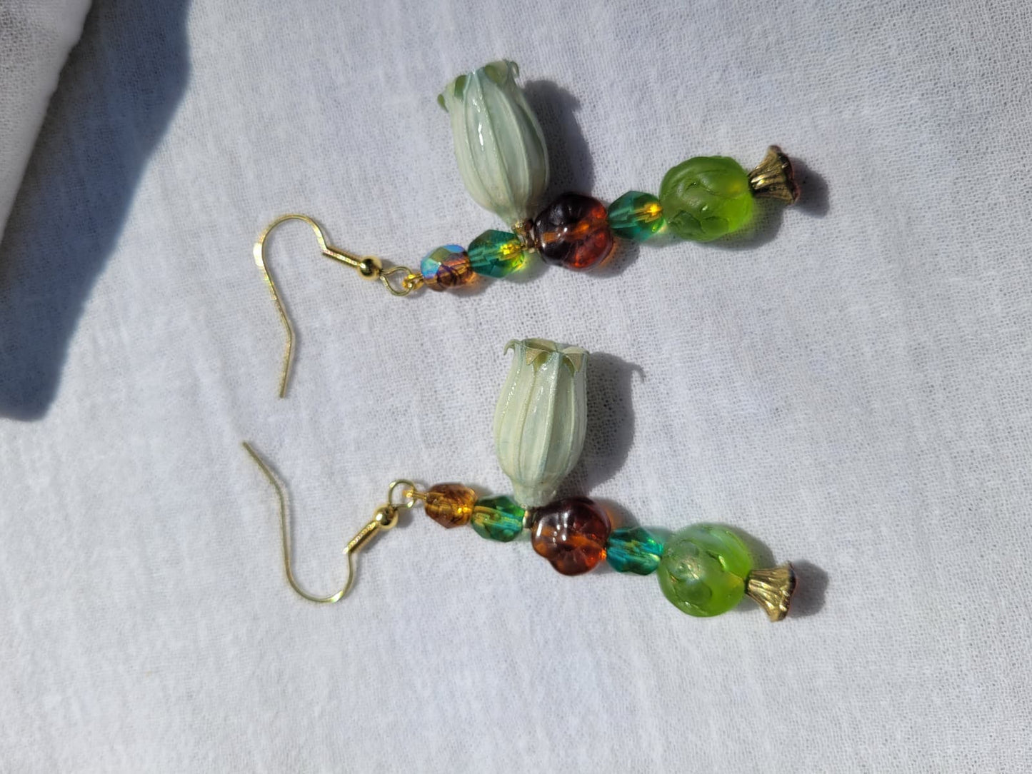 Beaded earrings with green and brown beads and natural dried flowers on a light fabric background; gold-coloured hooks.