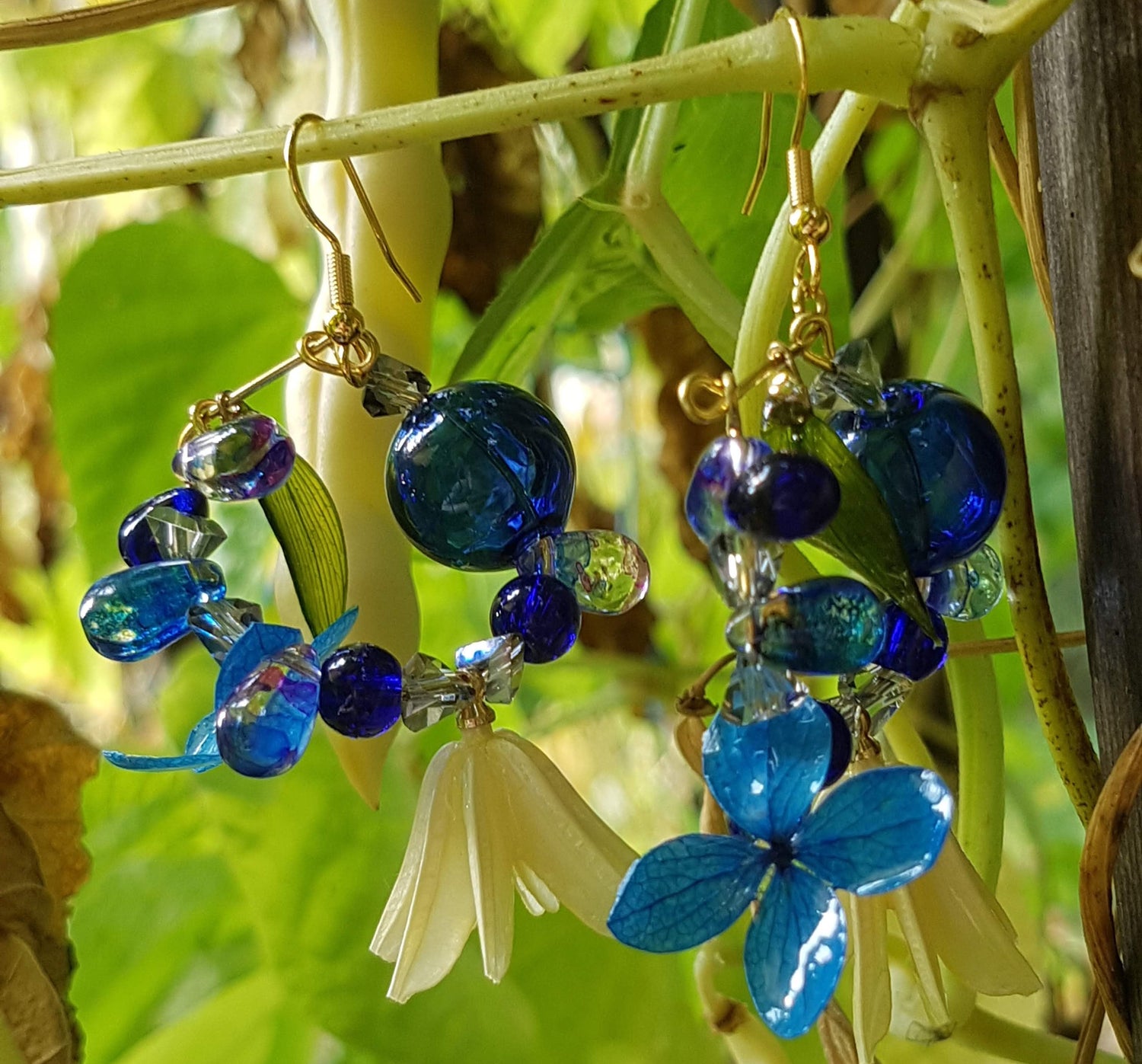 Blue floral earrings with gold accents hanging on green leaves.