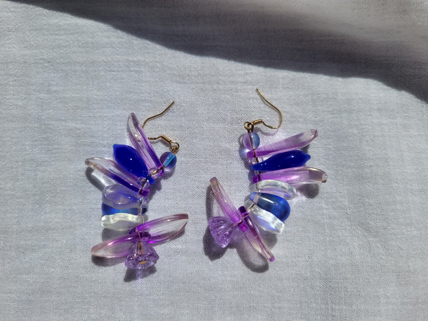 Colorful petal-shaped earrings with purple, blue and white glass beads on a light gray fabric background.