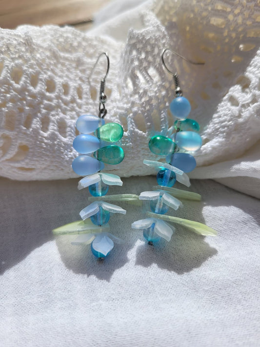 Pair of earrings with blue and green glass beads, acrylic leaves and small flowers on a white fabric background