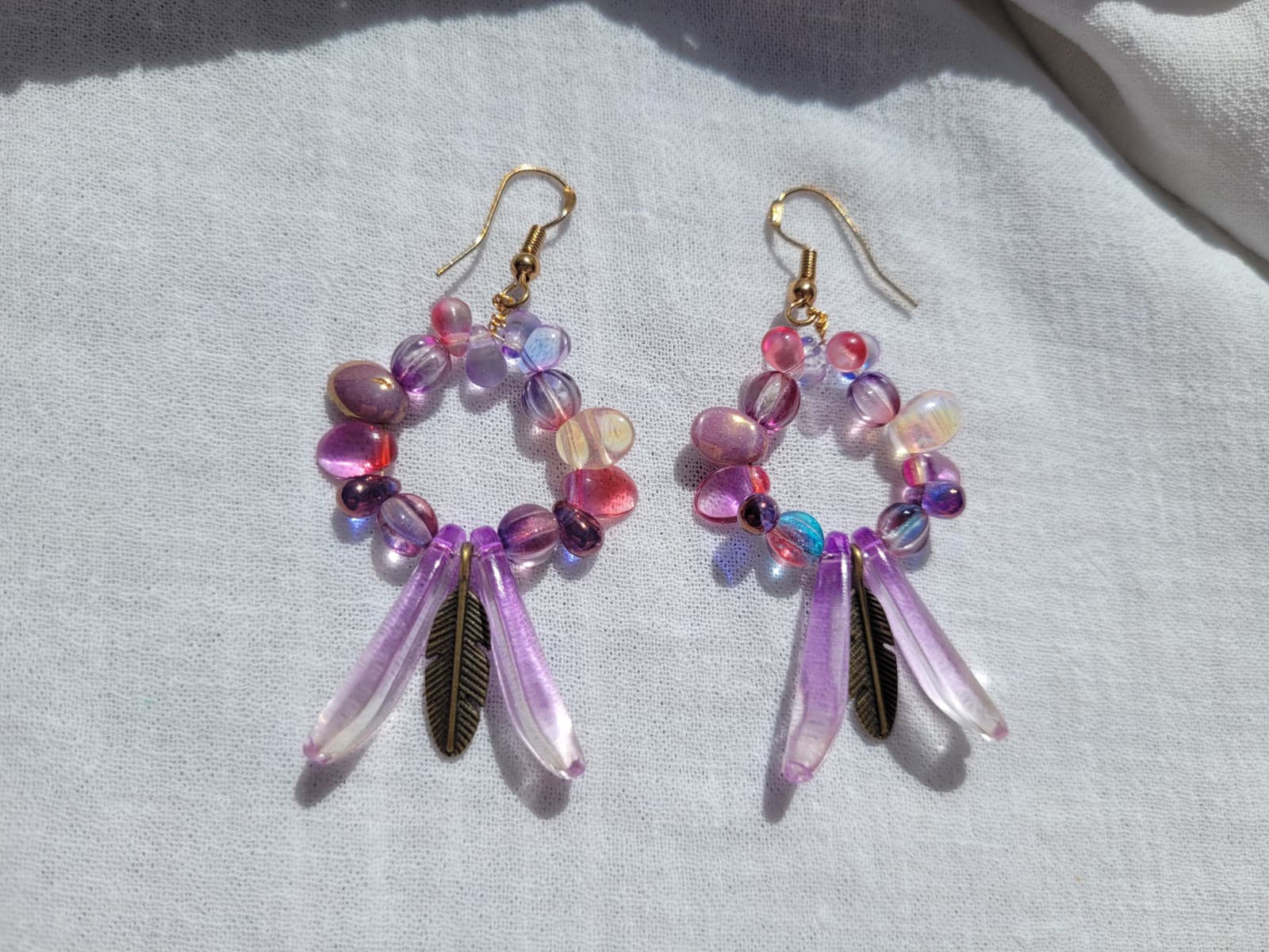 Pair of round dangle beaded earrings with pink and purple beads and bronze feather charms on a light gray background; gold-plated hooks