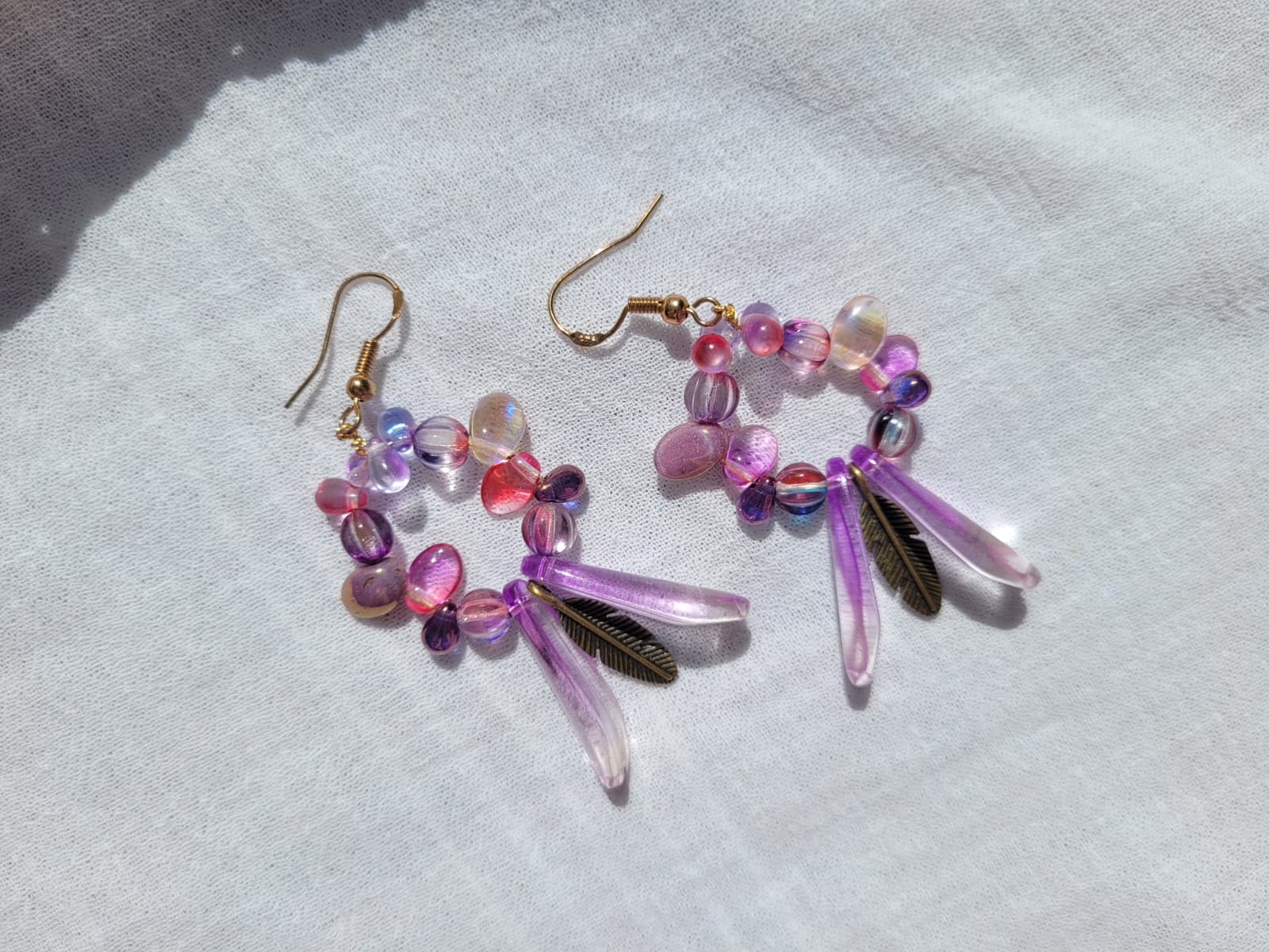 Pair of round dangle beaded earrings with pink and purple beads and bronze feather charms on a light gray background; gold-plated hooks