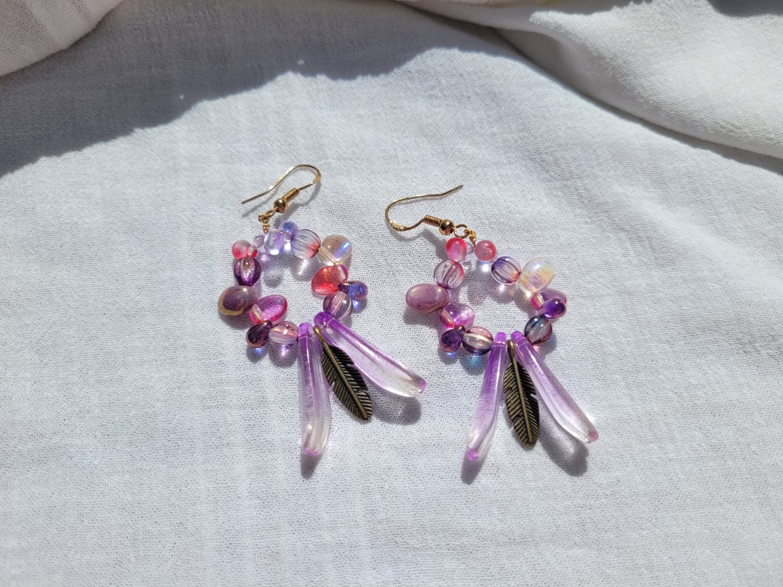 Pair of round dangle beaded earrings with pink and purple beads and bronze feather charms on a light gray background; gold-plated hooks