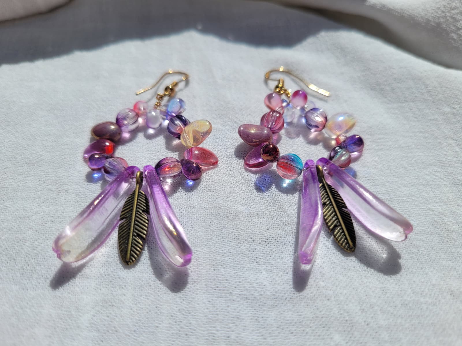 Pair of round dangle beaded earrings with pink and purple beads and bronze feather charms on a light gray background; gold-plated hooks
