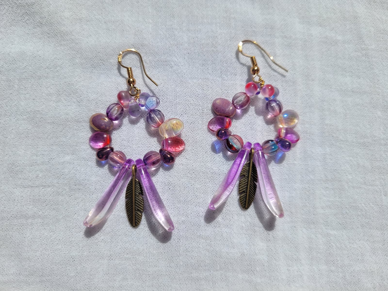 Pair of round dangle beaded earrings with pink and purple beads and bronze feather accent on a light gray background