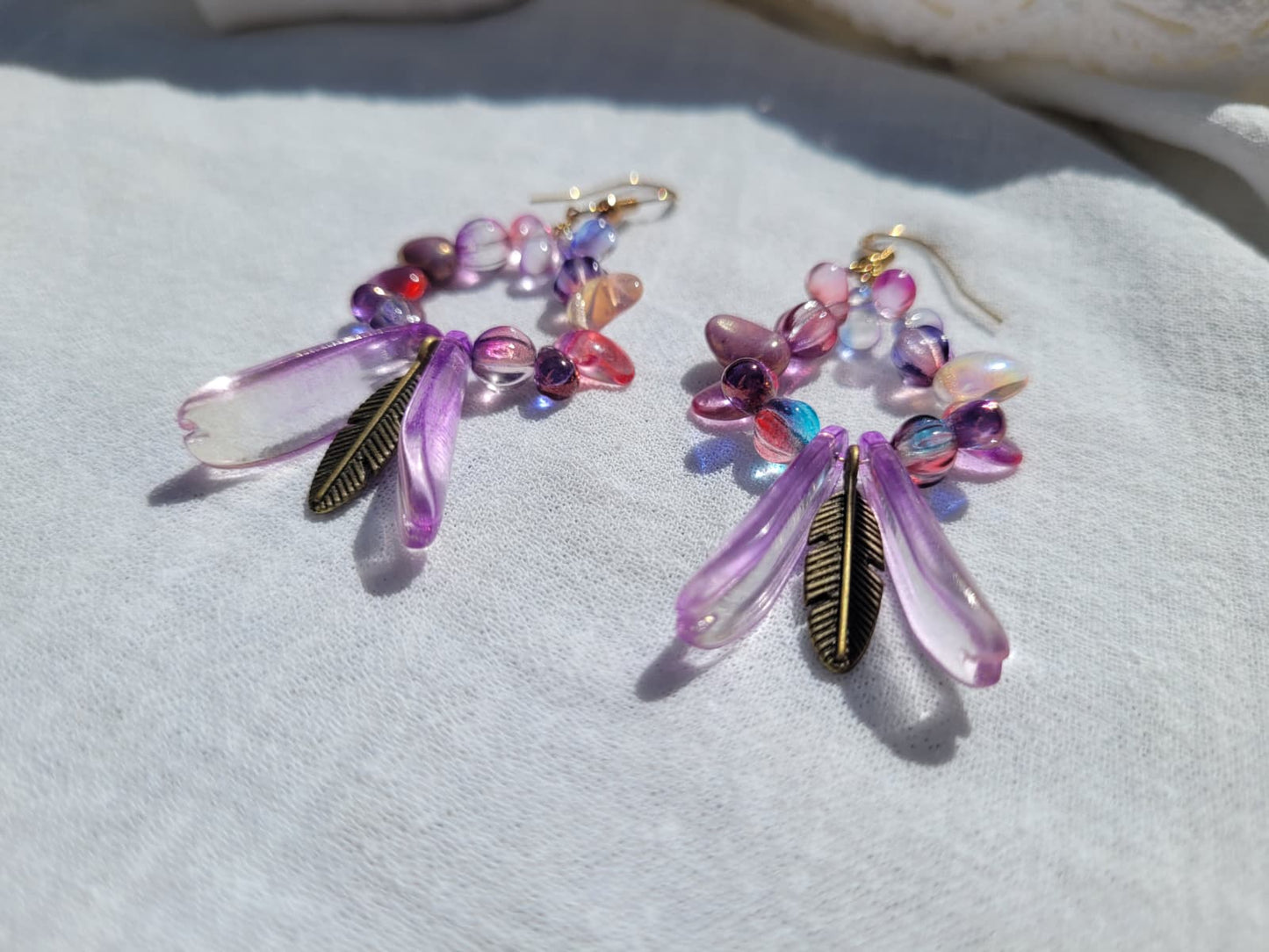 Pair of round dangle beaded earrings with pink and purple beads and bronze feather charms on a light gray background; gold-plated hooks