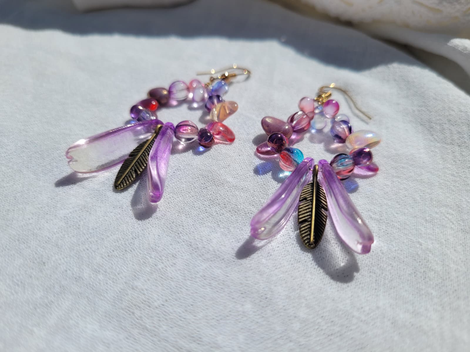 Pair of round dangle beaded earrings with pink and purple beads and bronze feather charms on a light gray background; gold-plated hooks