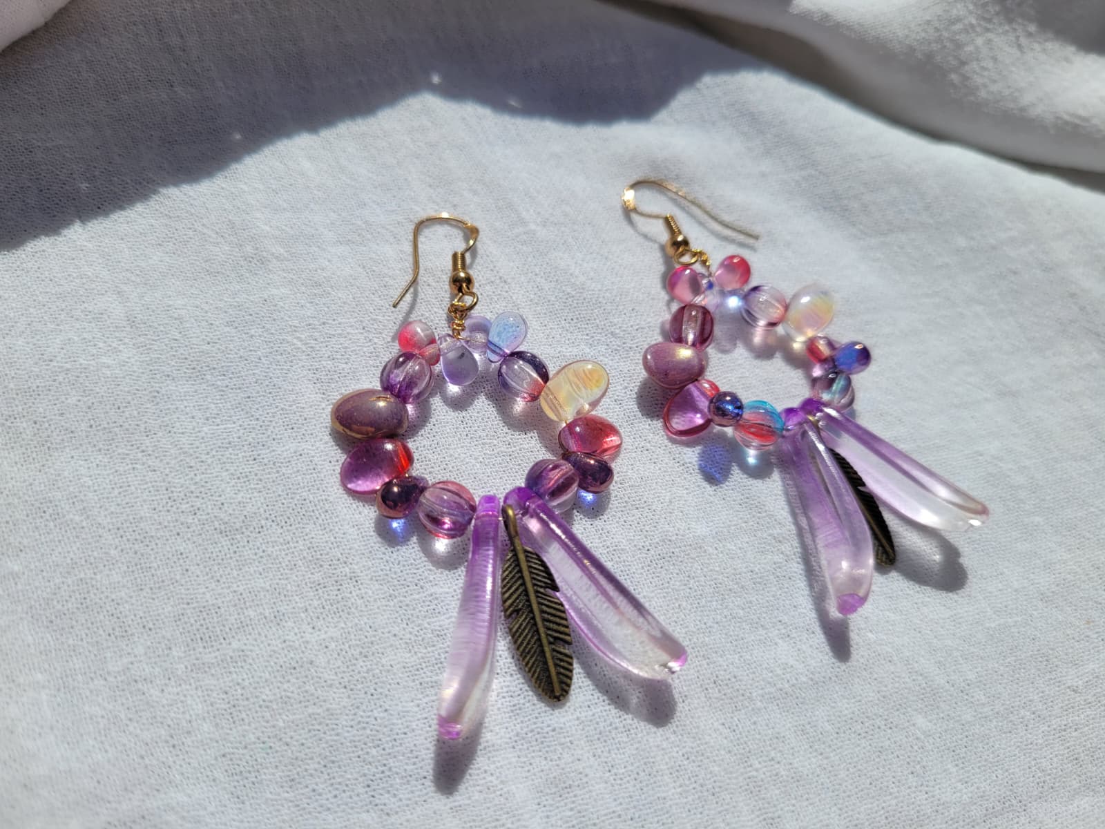 Pair of round dangle beaded earrings with pink and purple beads and bronze feather charms on a light gray background; gold-plated hooks