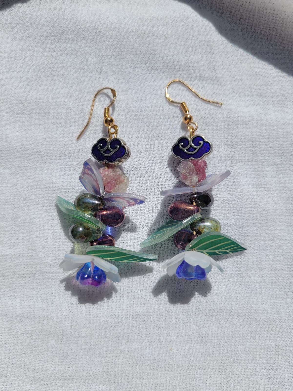 Colourful earrings with butterfly, floral and leaves details, with Cloisonne cloud-shaped beads on a light background