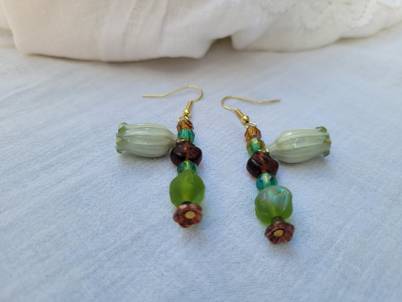 Beaded floral earrings with green and brown beads and natural dried flowers on a light fabric background; gold-coloured hooks.