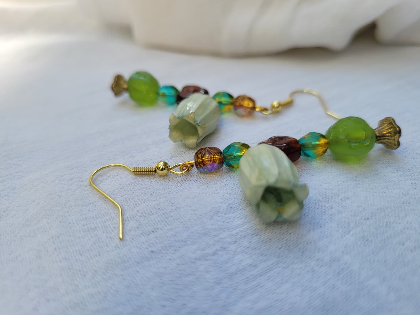 Beaded earrings with green and brown beads and natural dried flowers on a light fabric background; gold-coloured hooks.