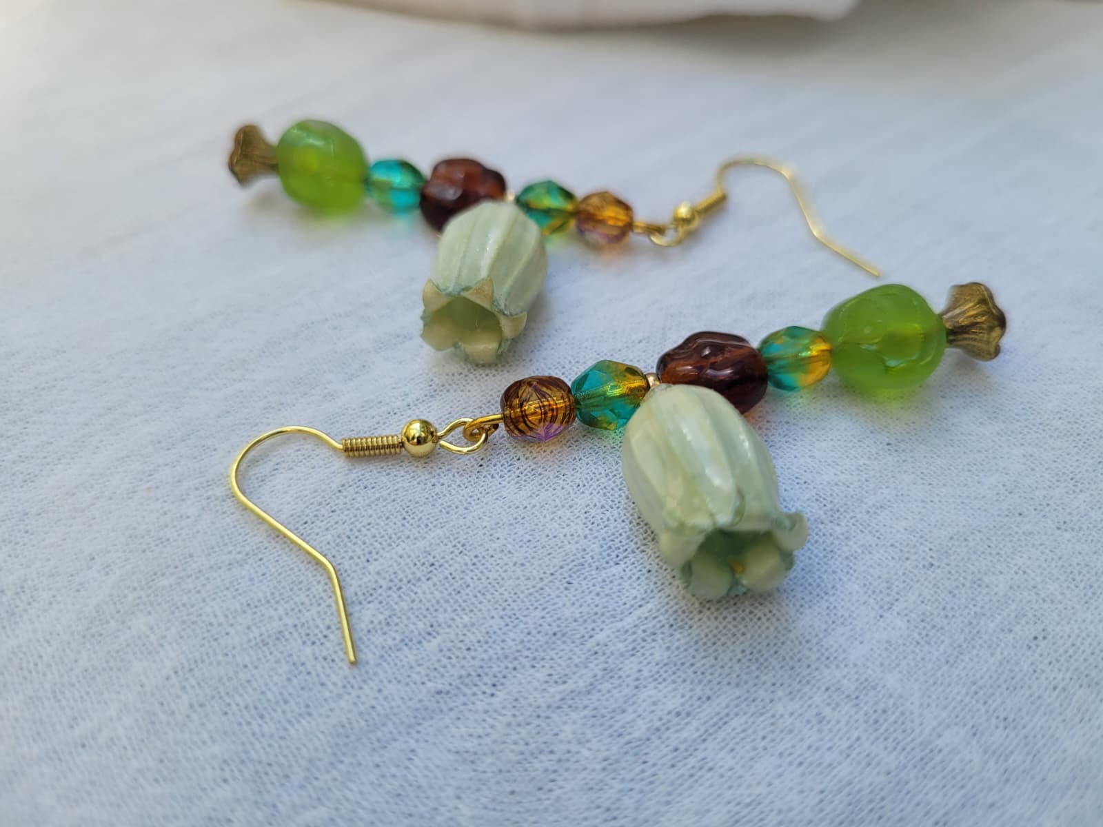 Beaded earrings with green and brown beads and natural dried flower on a light fabric background; gold-coloured hooks.