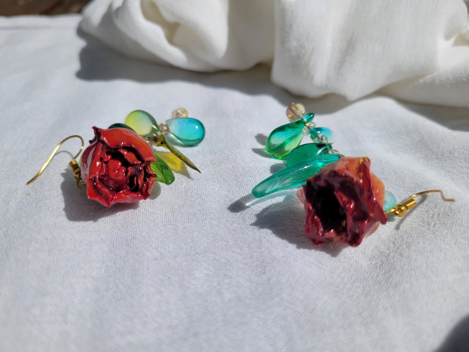Pair of floral earrings with real red rose buds and leaves, as well as green beads on a white fabric background