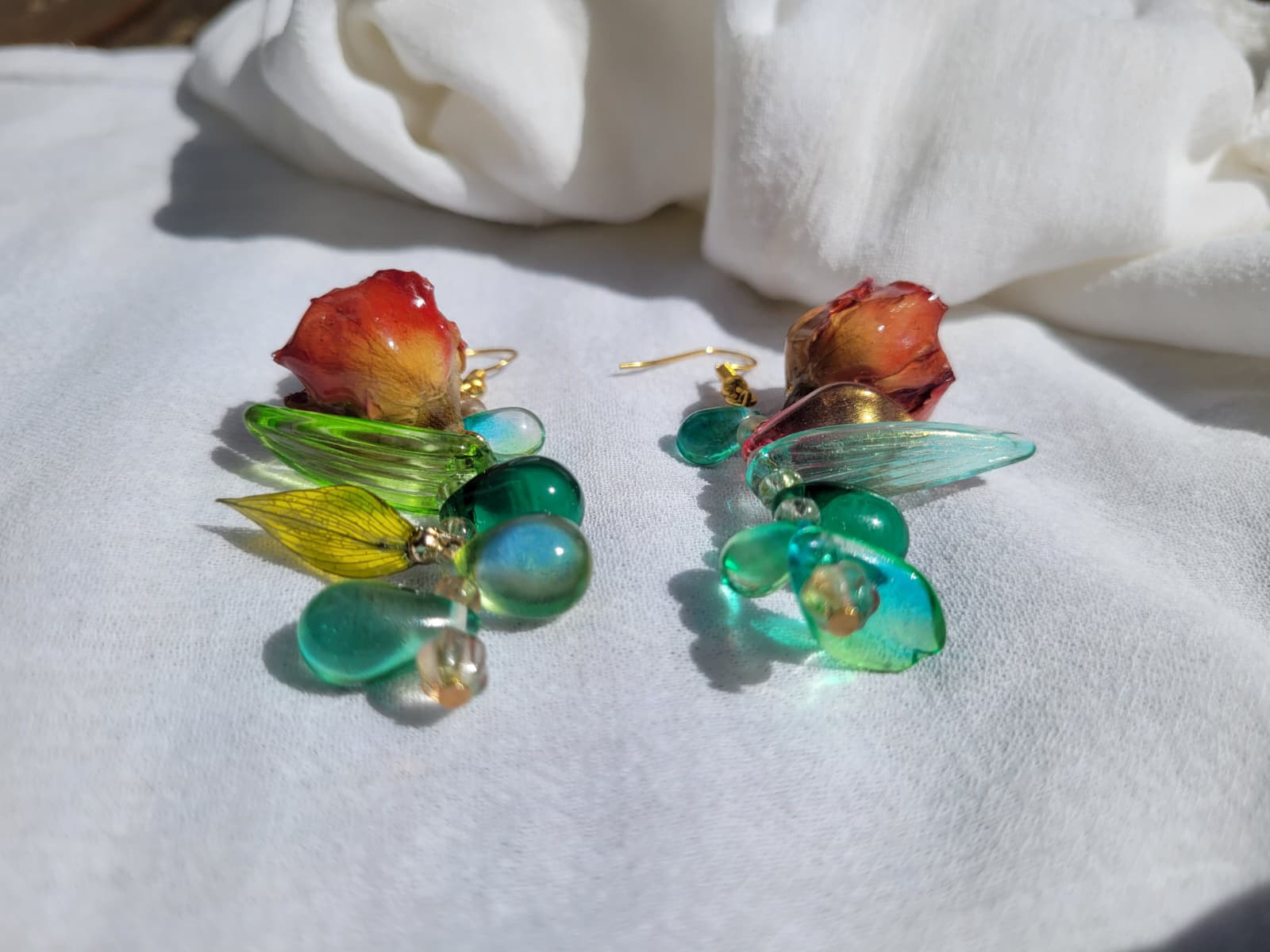 Pair of floral earrings with real red rose buds and leaves, as well as green beads on a white fabric background