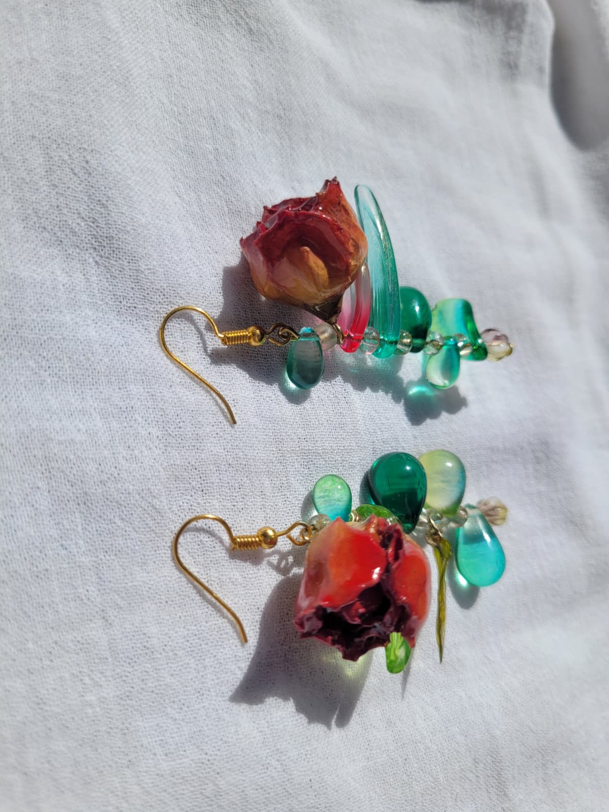 Pair of floral earrings with real red rose buds and leaves, as well as green beads on a white fabric background