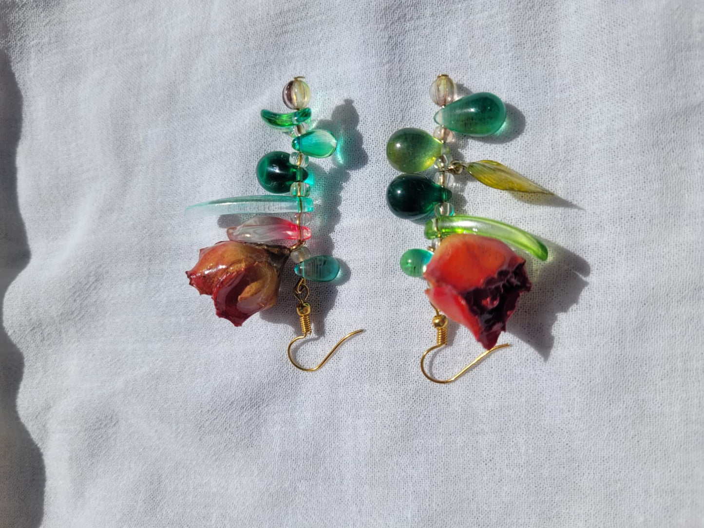Pair of floral earrings with real red rose buds and leaves, as well as green beads on a white fabric background