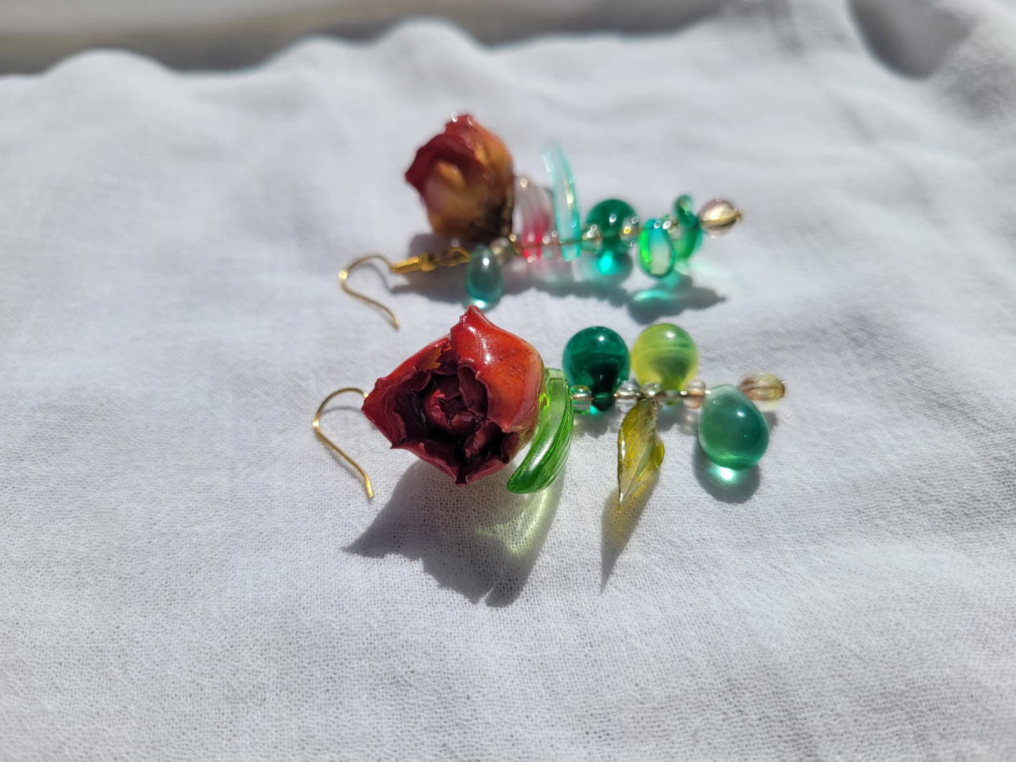 Pair of floral earrings with real red rose buds and leaves, as well as green beads on a white fabric background