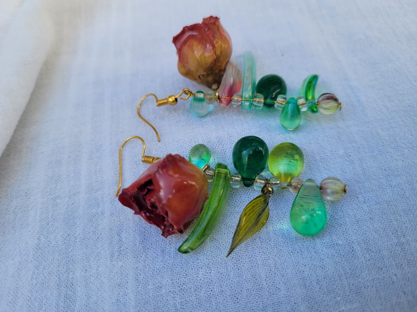 Pair of floral earrings with real red rose buds and leaves, as well as green beads on a white fabric background