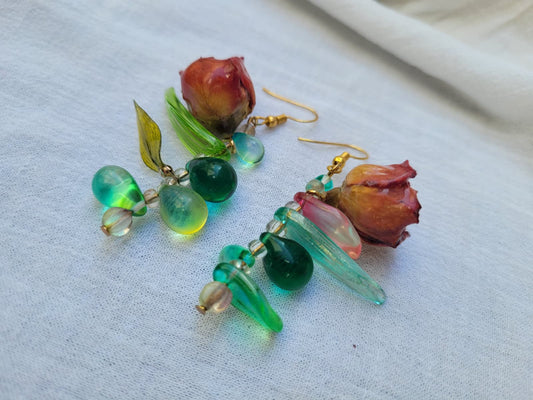 Pair of floral earrings with real red rose buds and leaves, as well as green beads on a white fabric background