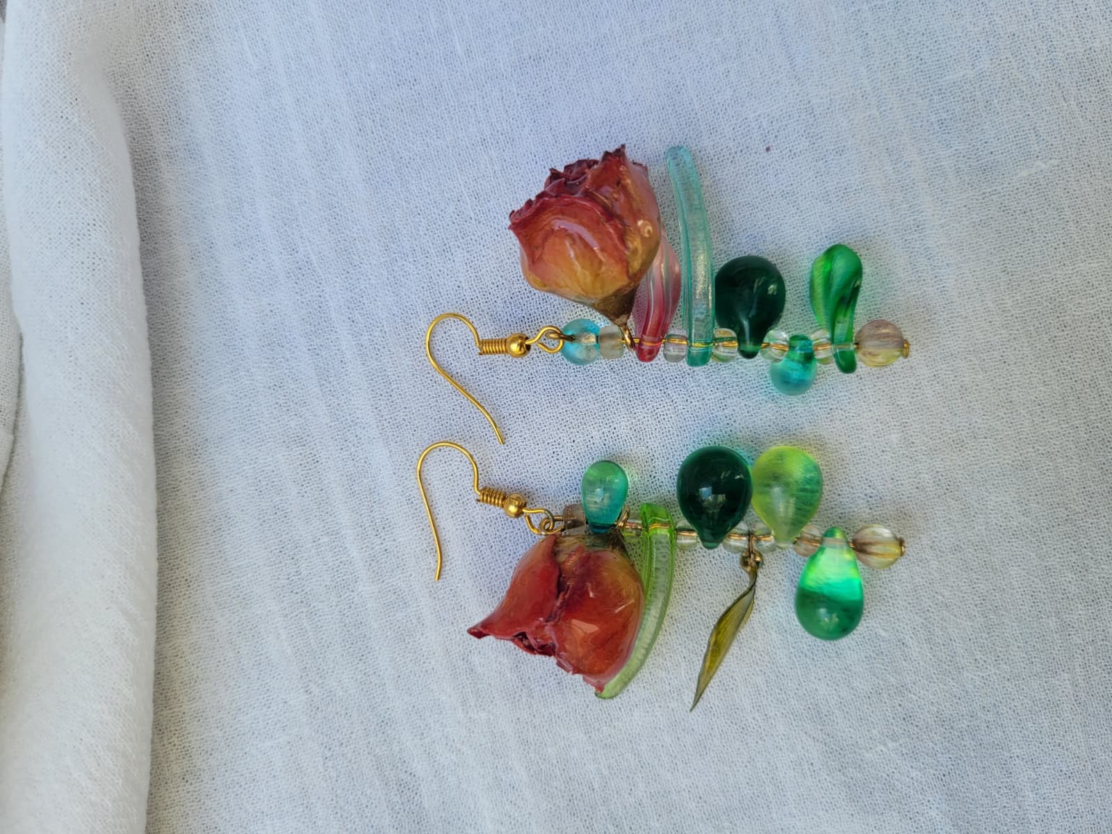 Pair of floral earrings with real red rose buds and leaves, as well as green beads on a white fabric background