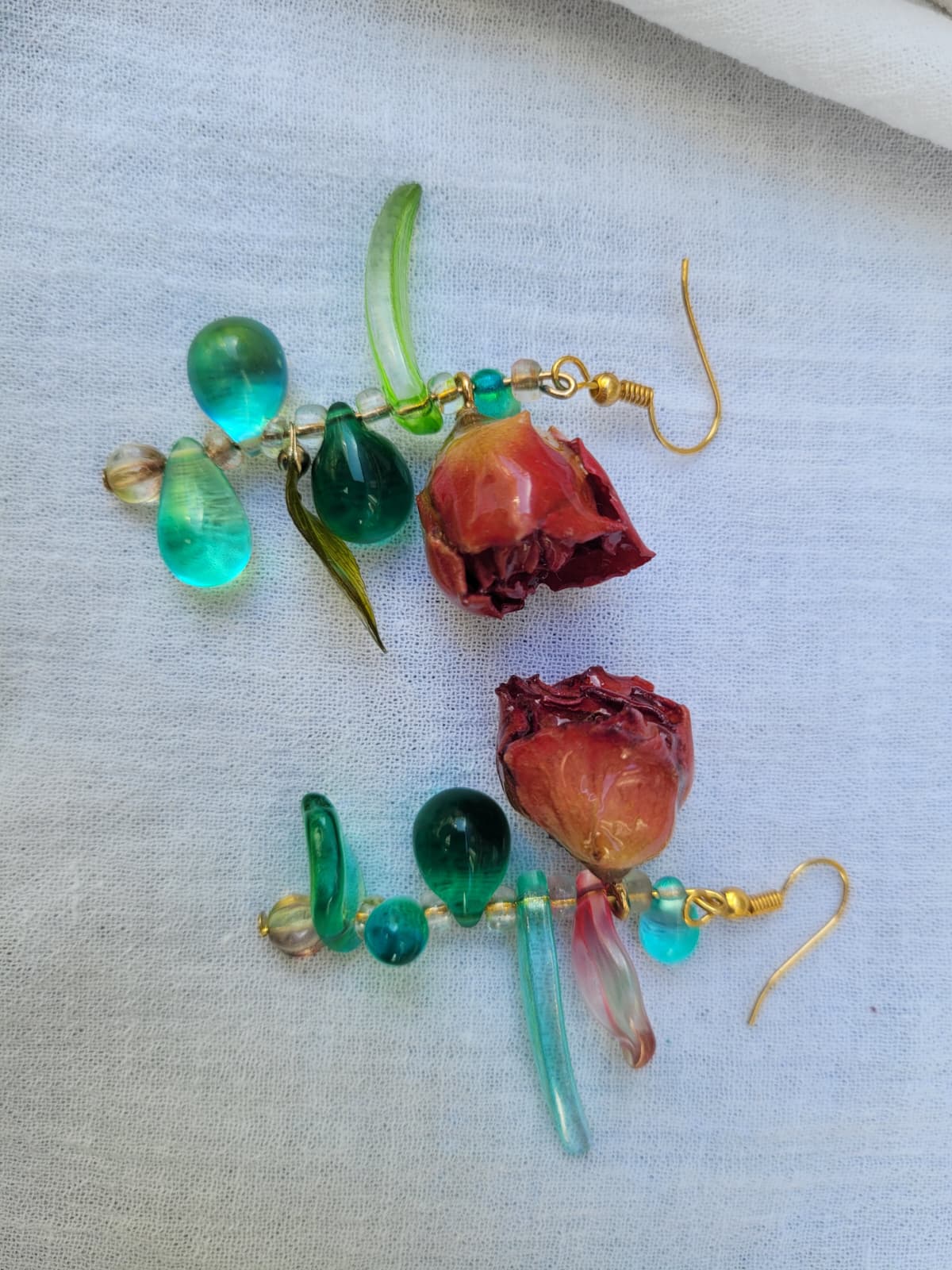 Pair of floral earrings with real red rose buds and leaves, as well as green beads on a white fabric background