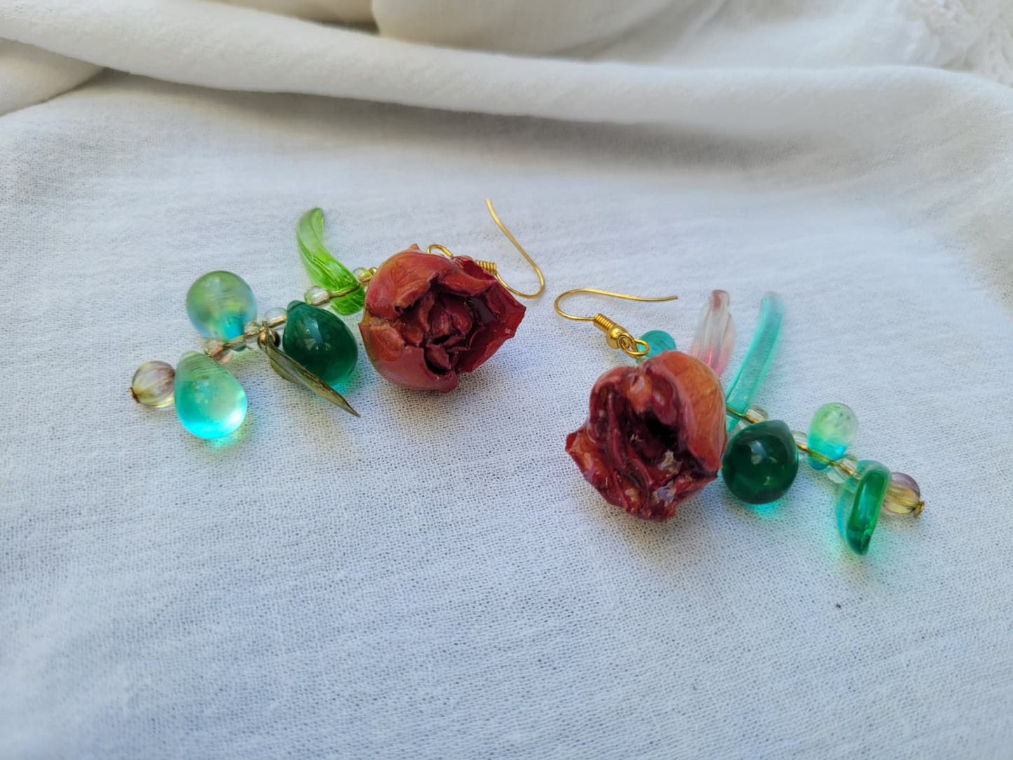 Pair of floral earrings with real red rose buds and leaves, as well as green beads on a white fabric background