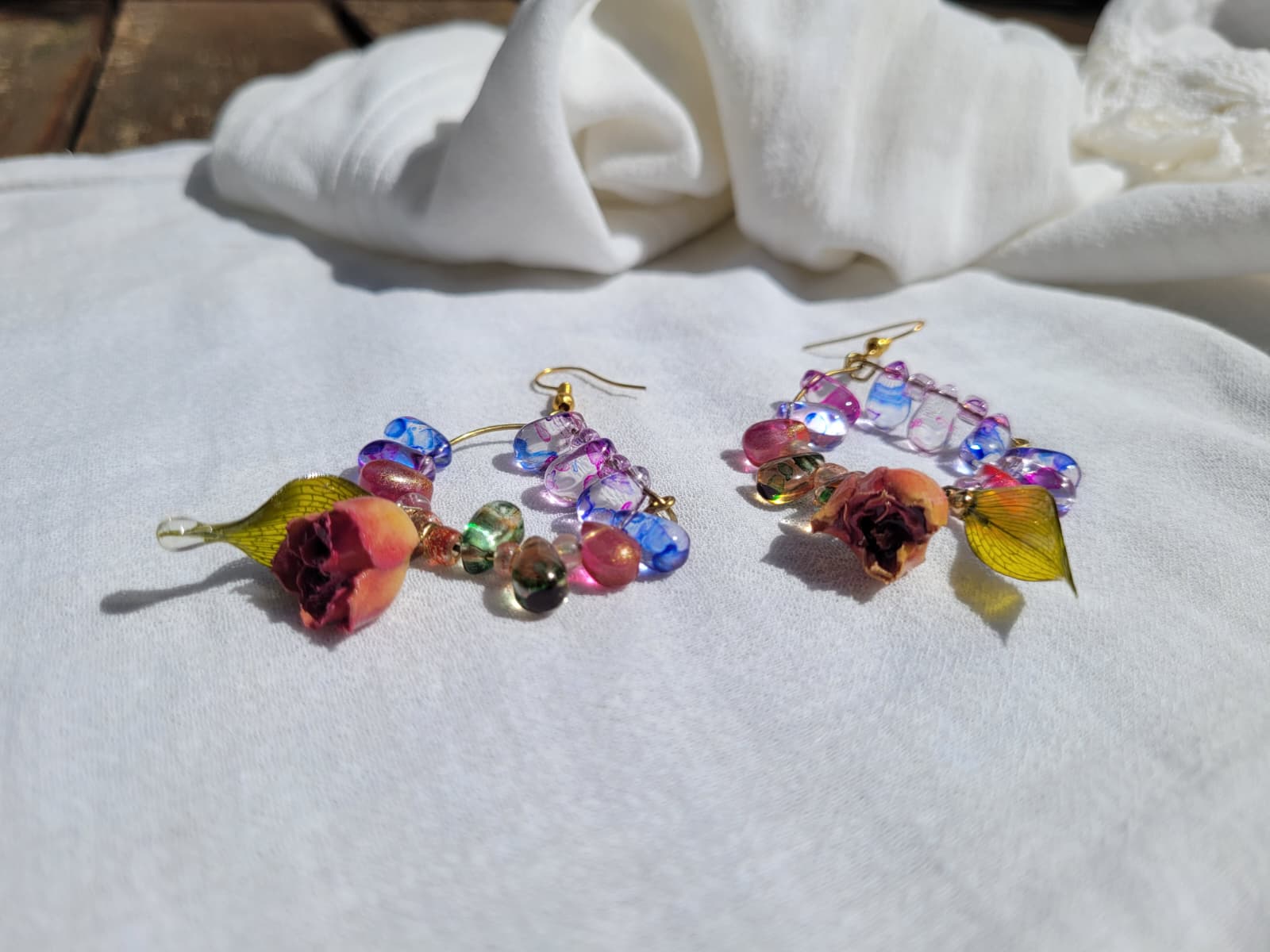 Colourful floral beaded earrings with natural dried leaves and yellow roses on a white fabric background; gold-coloured hooks