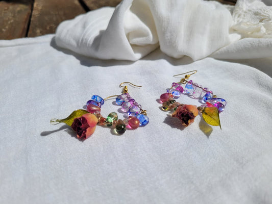 Colourful floral beaded earrings with natural dried leaves and yellow roses on a white fabric background; gold-coloured hooks