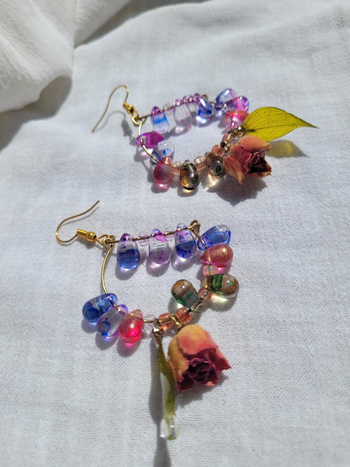 Colourful floral beaded earrings with natural dried leaves and yellow roses on a white fabric background; gold-coloured hooks