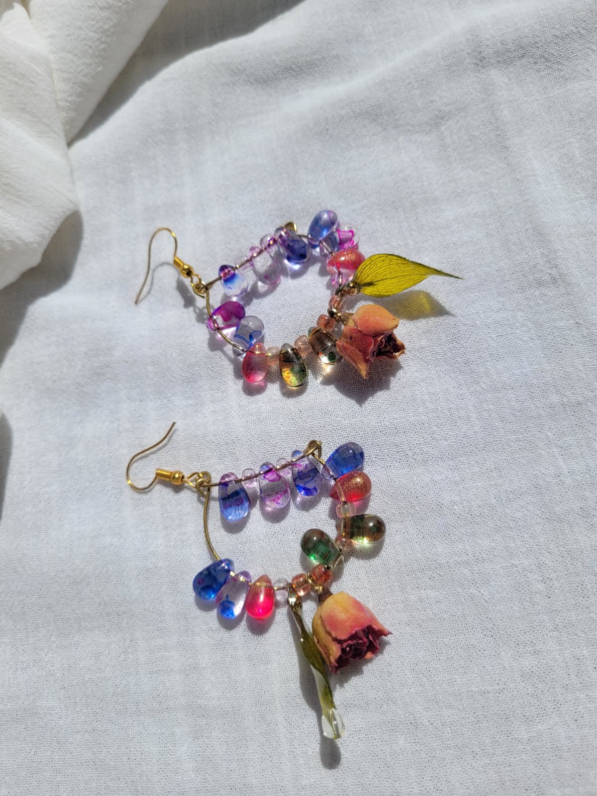 Colourful floral beaded earrings with natural dried leaves and yellow roses on a white fabric background; gold-coloured hooks