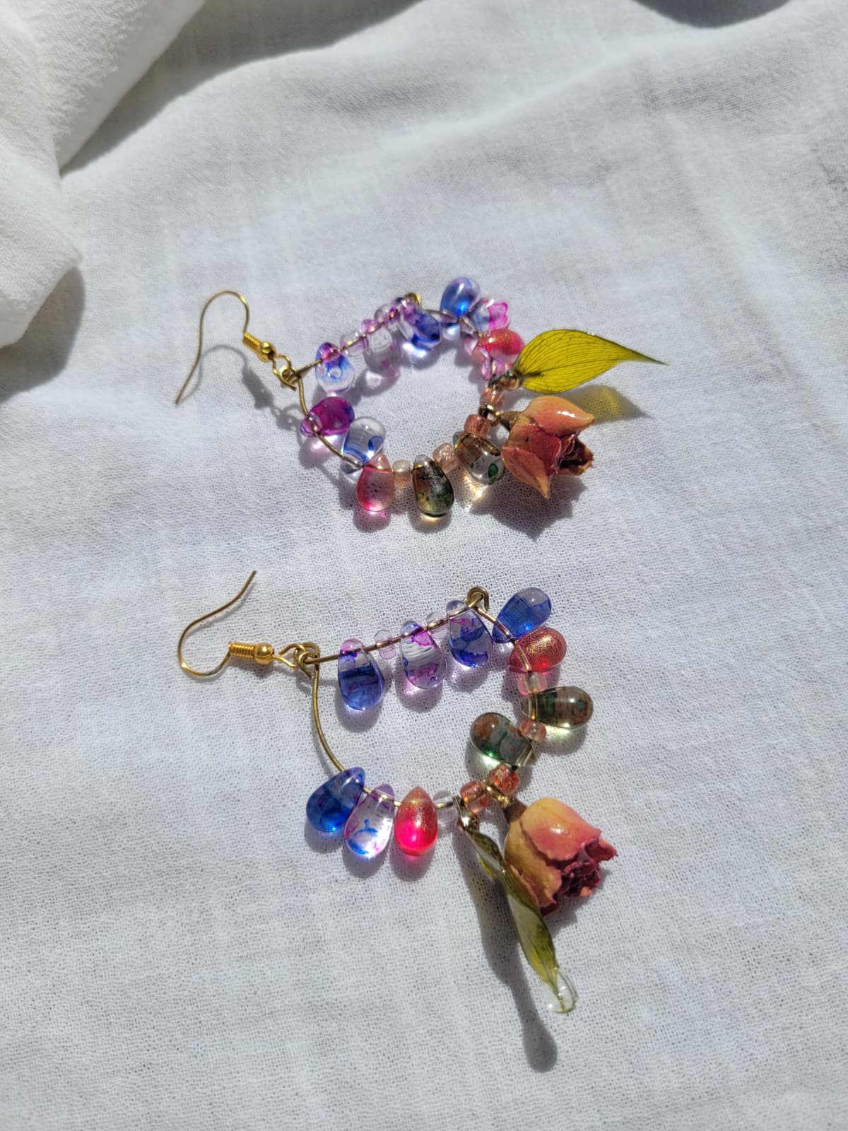 Colourful floral beaded earrings with natural dried leaves and yellow roses on a white fabric background; gold-coloured hooks