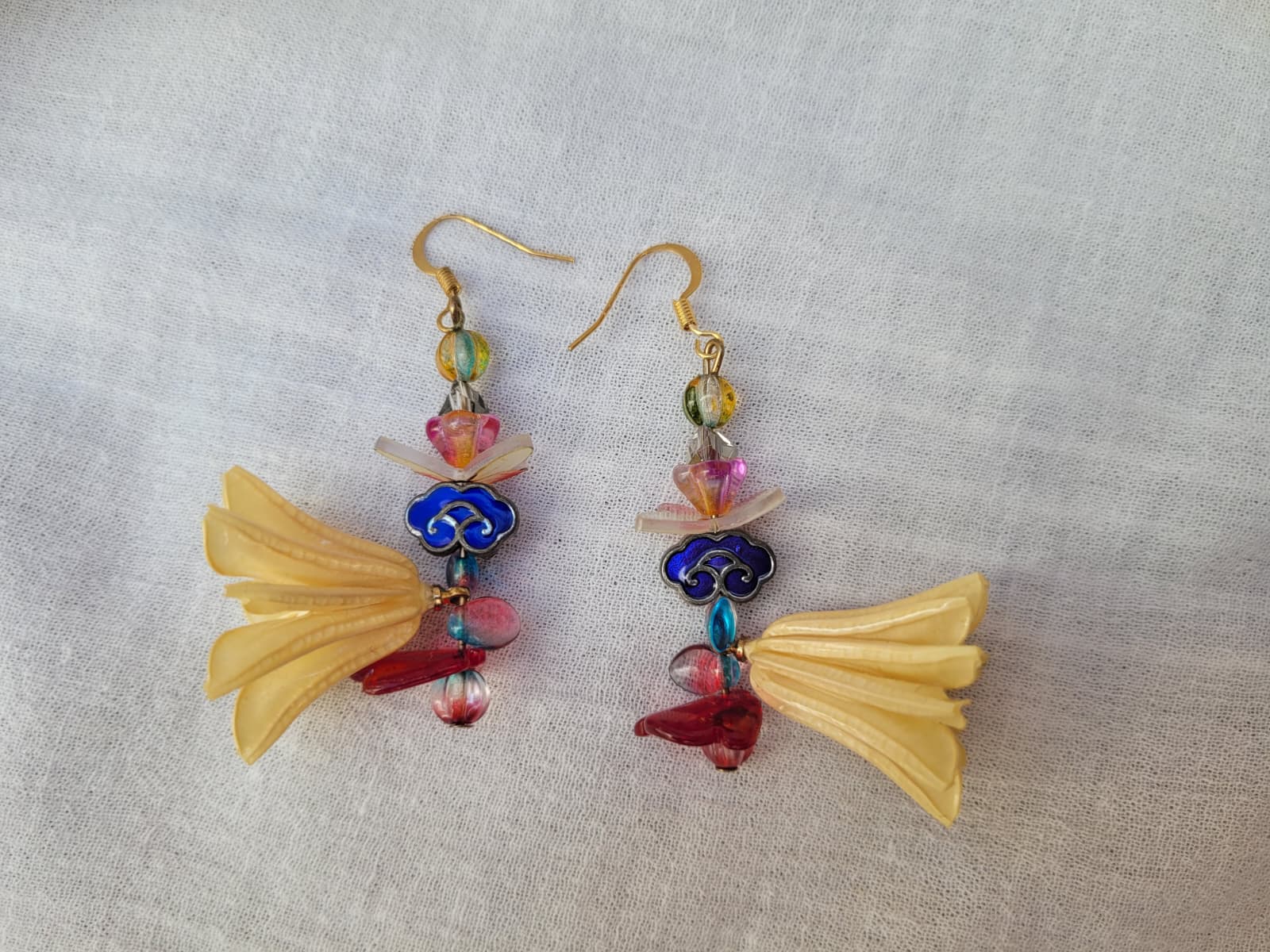 Colourful floral earrings with dried Nigella Flowers, cloud-shaped Cloisonne beads, butterflies and colourful glass beads on a light fabric background.
