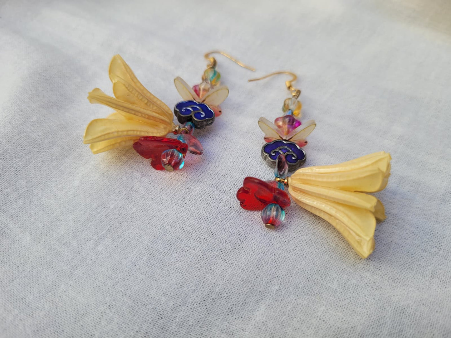 Colourful floral earrings with dried Nigella Flowers, cloud-shaped Cloisonne beads, butterflies and colourful glass beads on a light fabric background.