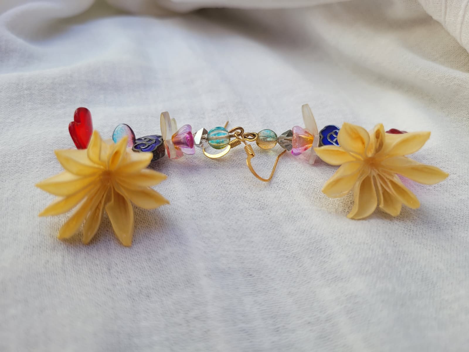 Colourful floral earrings with dried Nigella Flowers, cloud-shaped Cloisonne beads, butterflies and colourful glass beads on a light fabric background.