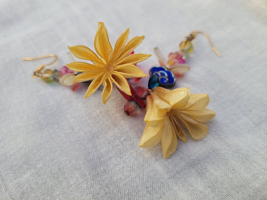 Colourful floral earrings with dried Nigella Flowers, cloud-shaped Cloisonne beads, butterflies and colourful glass beads on a light fabric background.