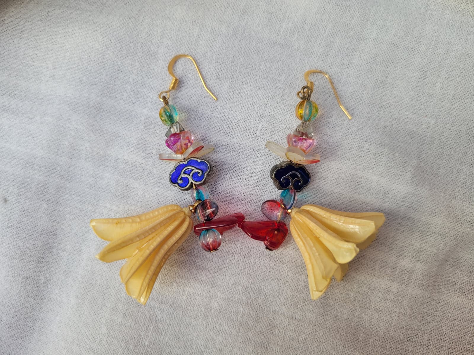 Colourful floral earrings with dried Nigella Flowers, cloud-shaped Cloisonne beads, butterflies and colourful glass beads on a light fabric background.