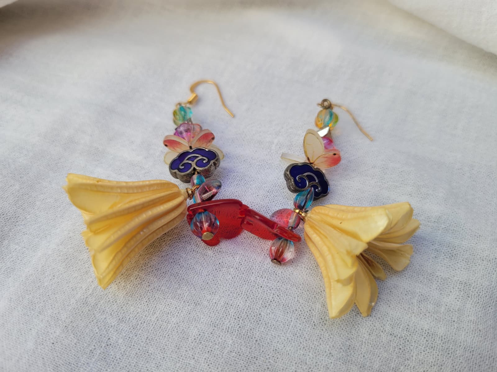 Colourful floral earrings with dried Nigella Flowers, cloud-shaped Cloisonne beads, butterflies and colourful glass beads on a light fabric background.