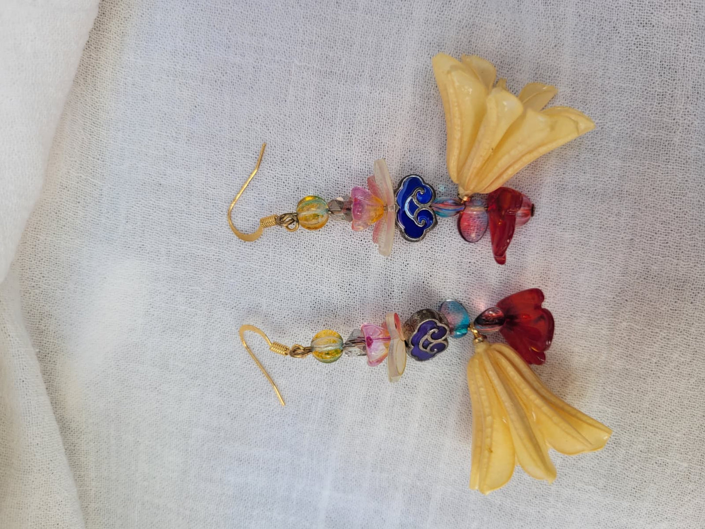 Colourful floral earrings with dried Nigella Flowers, cloud-shaped Cloisonne beads, butterflies and colourful glass beads on a light fabric background.