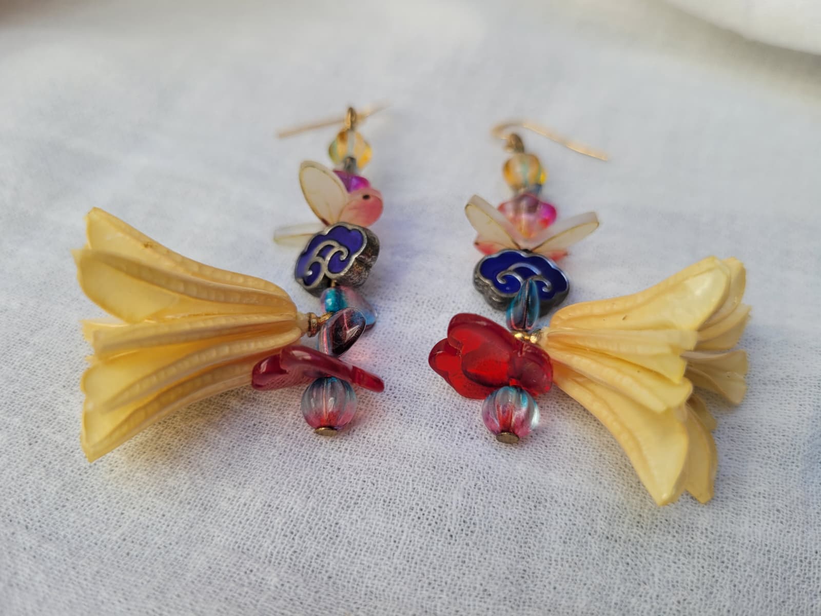 Colourful floral earrings with dried Nigella Flowers, cloud-shaped Cloisonne beads, butterflies and colourful glass beads on a light fabric background.
