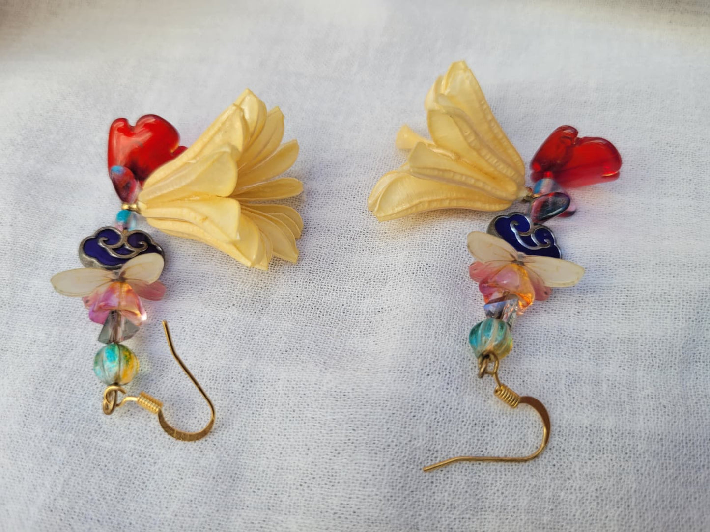 Colourful floral earrings with dried Nigella Flowers, cloud-shaped Cloisonne beads, butterflies and colourful glass beads on a light fabric background.