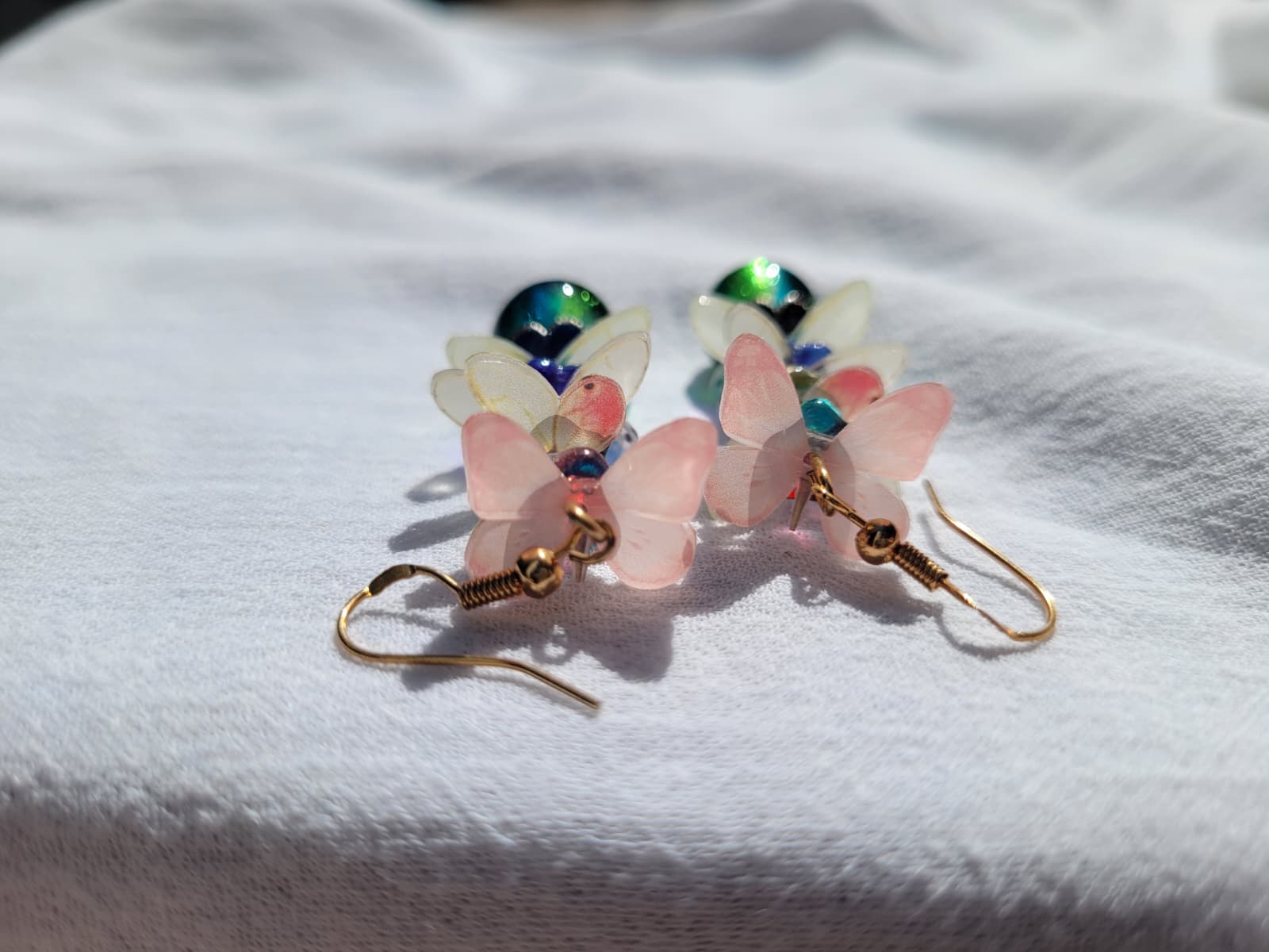 Butterfly-shaped earrings with pink wings and multicolored centers; gold plated hooks; on a white fabric background